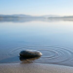 A minimalist, serene scene featuring a single smooth stone resting at the edge of a calm lake, creating gentle concentric ripples on the water’s surface under a clear sky.